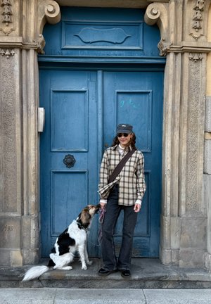 Joven mujer con camisa de cuadros y gorra, de pie sujetando con una correa a un perro blanco y negro sentado frente a una gran puerta de madera azul.
