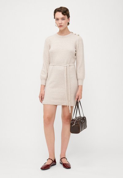 Vestido de punto beige con mangas largas, cintura ajustada con cinta y detalles de botones en el hombro. Combinado con un bolso marrón y zapatos burdeos.