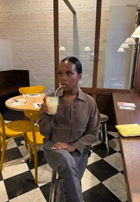 Frau mit dunklen Haaren und grauem Outfit, die auf einem Hocker sitzt und Limonade mit einem Strohhalm in einem Café mit gelben Stühlen und einem karierten Boden trinkt.