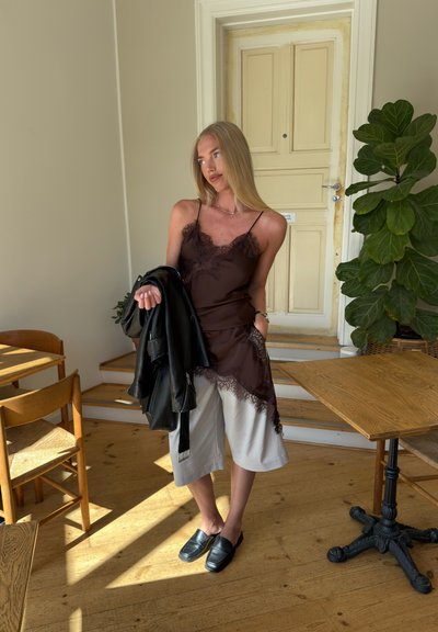 Camiseta de tirantes marrón con encaje sobre pantalones de pierna ancha blancos. Chaqueta de cuero negra sobre los hombros, con zapatos negros de slip-on. Entorno interior minimalista.