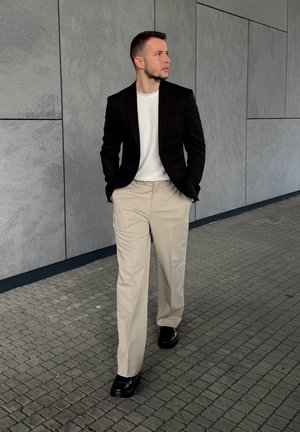Blazer negro sobre una camiseta blanca, pantalones beiges y zapatos negros; pose de pie contra una pared gris texturizada y un pavimento.