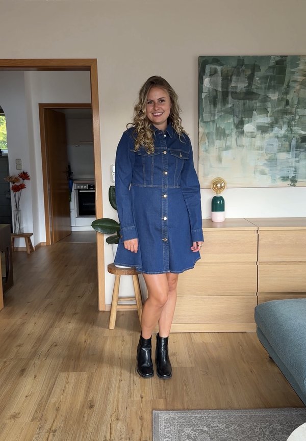 Mujer con pelo rizado que lleva un vestido vaquero azul y botas negras, de pie en una sala de estar moderna con suelos de madera y arte abstracto en la pared.