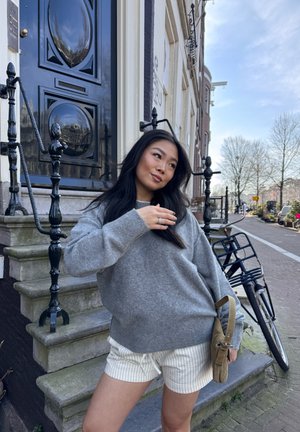 Suéter gris de gran tamaño, pantalones cortos blancos a rayas y una bolsa de pana marrón. Barandillas y escalones negros en una escena de calle con una bicicleta cercana.