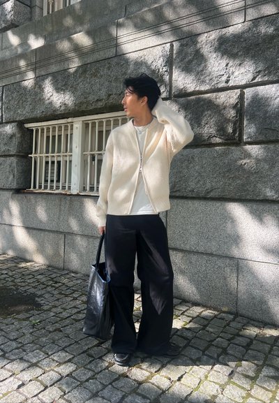 Cardigan de canalé crema con cremallera, llevado sobre una camisa blanca; combinado con pantalones anchos negros y una bolsa tote negra. Fondo de adoquines.
