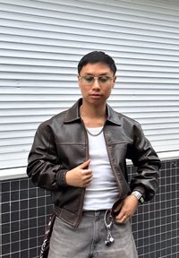 Brązowa skórzana kurtka bomberka z kołnierzem, zamkiem błyskawicznym i ściągaczami przy rękawach. Noszona na białym topie na ramiączkach, w połączeniu z szarymi jeansami i dodatkami.