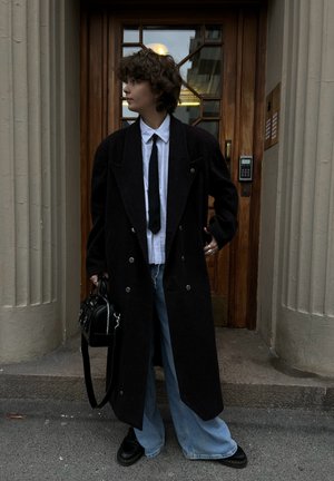 Abrigo largo negro de lana con cierre de doble botonadura, llevado sobre una camisa blanca y una corbata negra. Jeans de mezclilla azul sueltos y zapatos negros. Bolso negro.