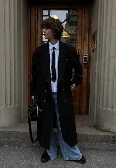 Abrigo largo negro de lana con cierre de doble botonadura, llevado sobre una camisa blanca y una corbata negra. Jeans de mezclilla azul sueltos y zapatos negros. Bolso negro.