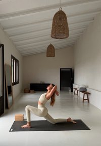 Femme en tenue de sport beige réalisant une posture de fente basse de yoga sur un tapis noir dans une pièce minimaliste avec un banc en bois, un miroir et des lampes en osier suspendues.
