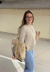 Beige strikket genser med ribbestrikket hals, lyseblå tettsittende jeans og en beige jakke i kunstpels drapert over den ene armen.