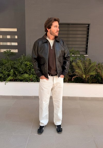 Hombre con cabello castaño que lleva una chaqueta de cuero negra, suéter oscuro, pantalones blancos y zapatillas negras, de pie con las manos en los bolsillos cerca de plantas.
