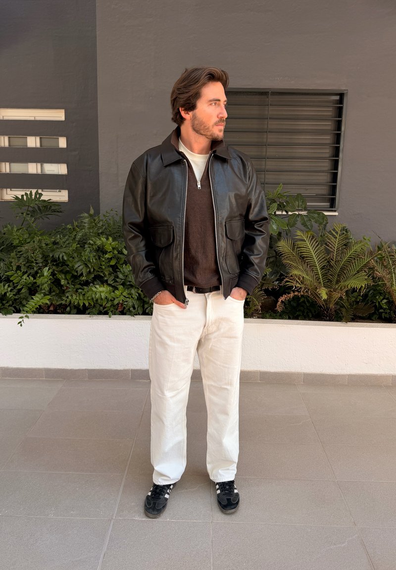 Barna hajú férfi fekete bőrdzsekiben, sötét pulóverben, fehér nadrágban és fekete tornacipőben, zsebre tett kézzel növények mellett áll.