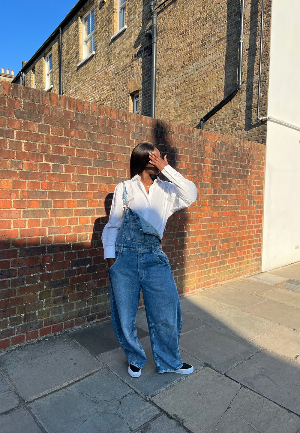 Svijetloplavi dječji kombinezon širokih nogavica, uparen s dugim bijelim majicom i crnim tenisicama, na pozadini od crvene cigle.