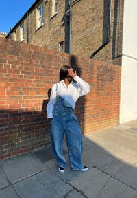 Svijetloplavi dječji kombinezon širokih nogavica, uparen s dugim bijelim majicom i crnim tenisicama, na pozadini od crvene cigle.