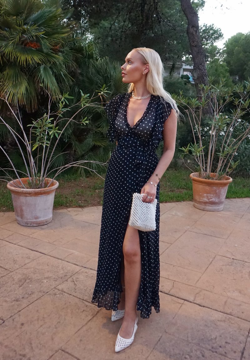Langes, navyblaues Kleid mit Punkten, rüschierten Ärmeln und einem seitlichen Schlitz, kombiniert mit weißen spitzen Absatzschuhen und einer strukturierten weißen Clutch.