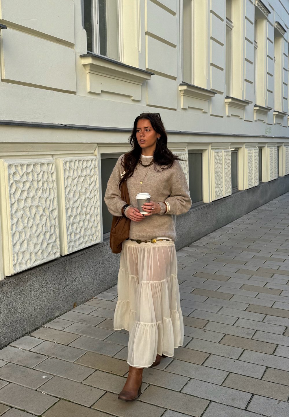 Pulover bej tricotat, fustă crem transparentă în straturi, cizme maro până la gleznă. Ține o ceașcă de cafea, completată cu o geantă maro și coliere suprapuse.