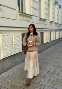 Pulover bej tricotat, fustă crem transparentă în straturi, cizme maro până la gleznă. Ține o ceașcă de cafea, completată cu o geantă maro și coliere suprapuse.