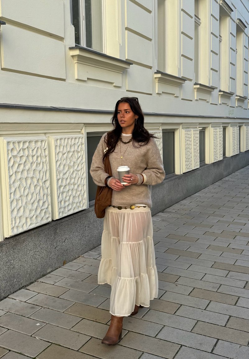 Suéter bege de malha, saia creme translúcida em camadas, botas castanhas até ao tornozelo. Segurando uma chávena de café, complementada com uma bolsa castanha e colares em camadas.