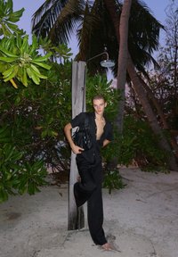 Camisa preta com botões e gola aberta, calças largas pretas e uma bolsa preta. O fundo apresenta folhagem tropical e um poste de madeira.