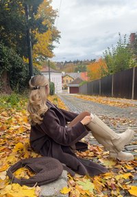 Brun lang frakke, beige knælange støvler, siddende på gule efterårsblade langs en brostensbelagt gade flankeret af farverige træer og bygninger.