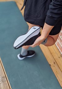 Chaussure de sport noire avec une tige texturée et une semelle en caoutchouc blanche, assortie à des chaussettes grises, posée sur un tapis d'exercice vert.