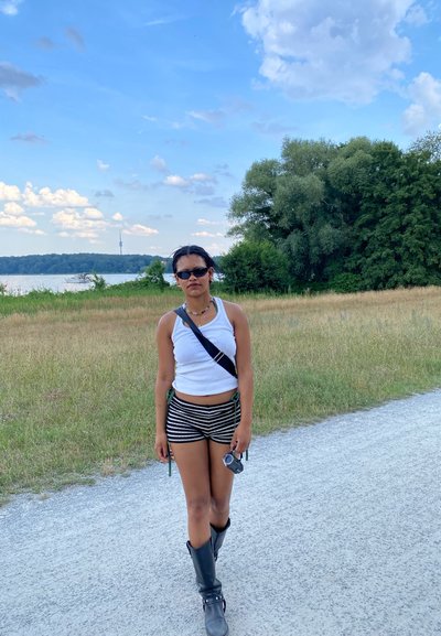 Una persona está de pie en un camino de grava, vistiendo una camiseta sin mangas blanca, pantalones cortos a rayas en blanco y negro, y botas negras hasta la rodilla, sosteniendo una cámara.