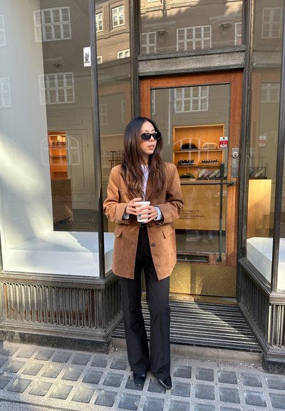 Blazer de ante marrón sobre una blusa blanca, combinado con pantalones acampanados negros y zapatos de punta. Sostiene una taza blanca, de pie frente a una entrada de cristal.