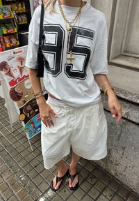 Fehér, oversized póló fekete "95" nyomattal, fehér bermuda rövidnadrággal, fekete papuccsal és zöld árnyalatú, rétegelt arany nyakláncokkal párosítva.