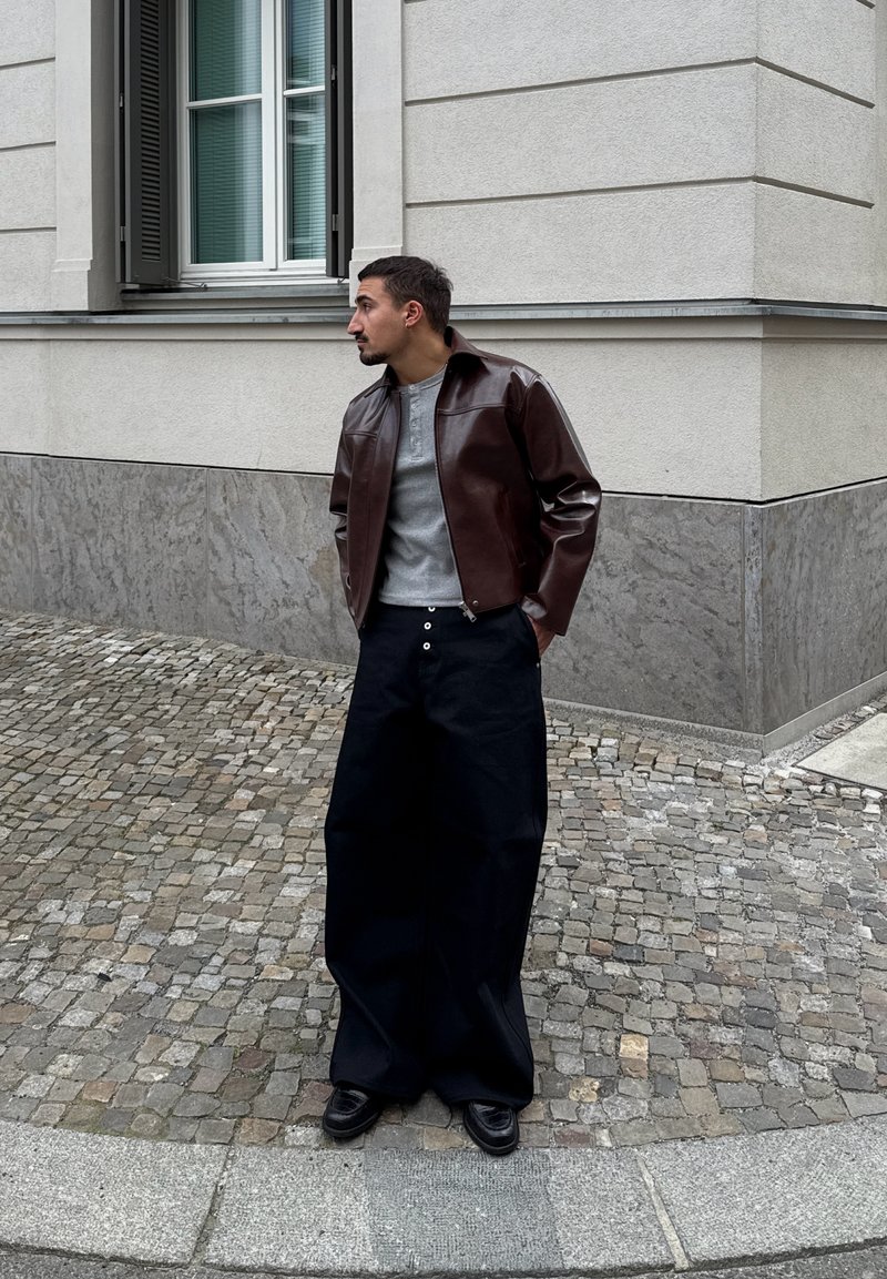 Homme portant une veste en cuir marron, une chemise grise et un pantalon noir large, debout dans une rue pavée près d’un bâtiment d’angle.