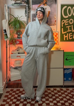 Sudadera con capucha gris con cremallera y pantalones deportivos a juego, hechos de material suave; combinados con un gorro negro y blanco. El fondo incluye varios zapatos y adornos.