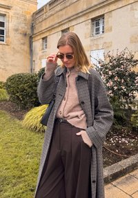 Cappotto grigio a quadri sopra un maglione di lana beige, abbinato a pantaloni larghi marroni. Accessoriato con occhiali da sole e una borsa nera.