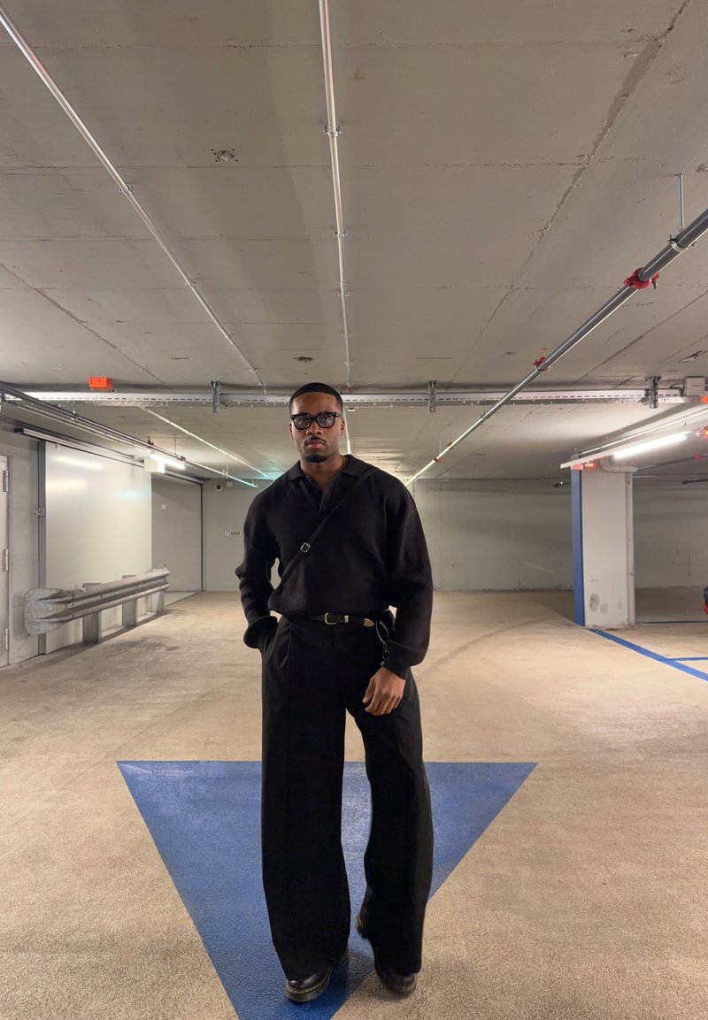 Black oversized shirt with long sleeves, paired with wide-leg trousers, both in black. Features a belt and glasses, set against a car park backdrop.