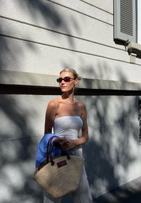 Saco de palha trançada com alças de couro castanho, uma blusa branca sem alças e calças de cor clara, complementado por uma camisa azul pendurada no braço.
