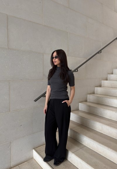 Mujer con cabello largo y oscuro, que lleva gafas de sol negras, camiseta gris oscuro y pantalones sueltos negros, está junto a unas escaleras de piedra beige con pasamanos metálico.
