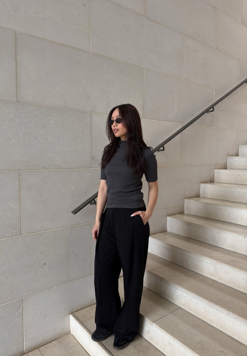 Femme aux longs cheveux foncés portant des lunettes de soleil noires, un haut gris foncé et un pantalon noir ample, debout près d'un escalier en pierre beige avec une rampe en métal.