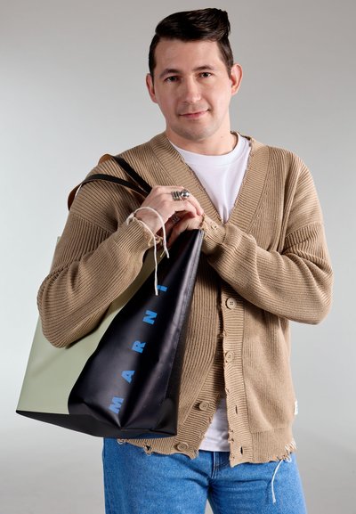 Cárdigan marrón de punto con cuello en V y botones visibles, combinado con jeans azules. Sosteniendo una bolsa de compra negra y verde con letras azules.