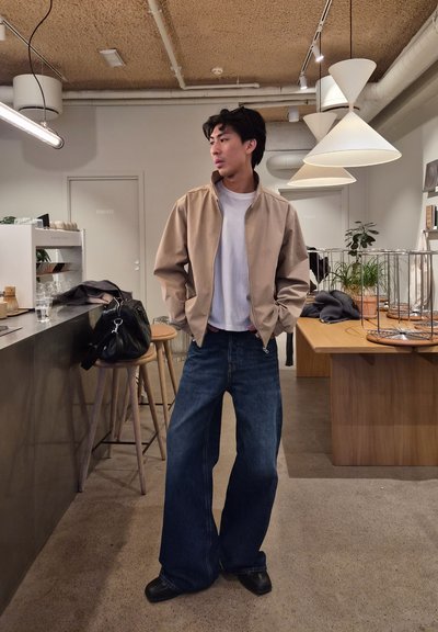 Hombre con chaqueta beige y vaqueros anchos de pie en una cafetería moderna con taburetes de madera, mesas, lámparas colgantes y plantas de interior.