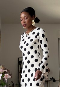 Langemouwjurk in het wit met grote zwarte stippen, met een ronde halslijn en een getailleerde pasvorm. Gouden accessoires en gestructureerde oorbellen.