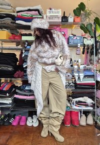 Personne portant une veste et un chapeau en fausse fourrure tachetée, un pantalon beige et des bottes souples, debout devant des étagères remplies de vêtements pliés et de chaussures.