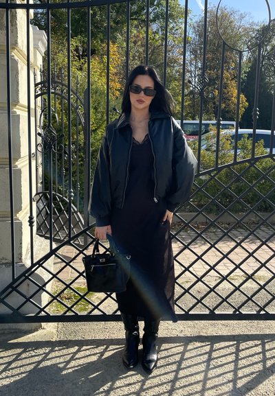 Chaqueta bomber negra con cuello acanalado, superpuesta sobre un vestido negro, combinada con botas negras hasta la rodilla y un pequeño bolso.