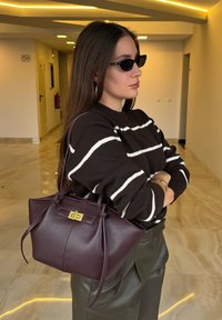 Sac en cuir bordeaux avec une texture lisse, doté d'un fermoir en laiton doré et de deux liens décoratifs ; associé à un pull rayé noir et blanc.