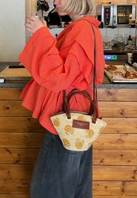 Πορτοκαλί υπερμεγέθη μπλούζα, σκούρα παντελόνια και τσάντα από ψάθα με κίτρινα λουλούδια και καφέ δερμάτινες λαβές.