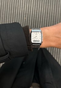 Ceas quartz Casio argintiu cu un cadran alb rectangular, bandă metalică și afișaj digital, așezat pe un fundal texturat întunecat.