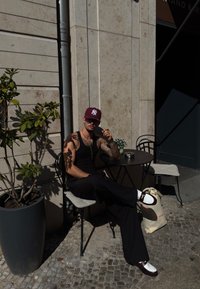 Črne široke hlače, črn top brez rokavov, burgundska kapa in bele čevlje z rjavimi poudarki. Sedi za majhno mizo blizu cvetlične rože.