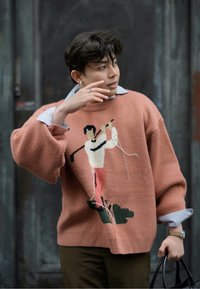 Rūsaina krāsā adīta džemperis ar izšūtu golfa spēlētāju dizainu. Tam ir brīva piegriezuma forma, ribotās piedurknes un zem tā ir slāņots gaismas zils krekls.