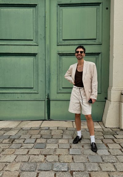 Blazer de lino de color beige claro, top marrón, pantalones cortos blancos con cordón, mocasines negros, calcetines blancos; de pie frente a puertas de madera verdes.