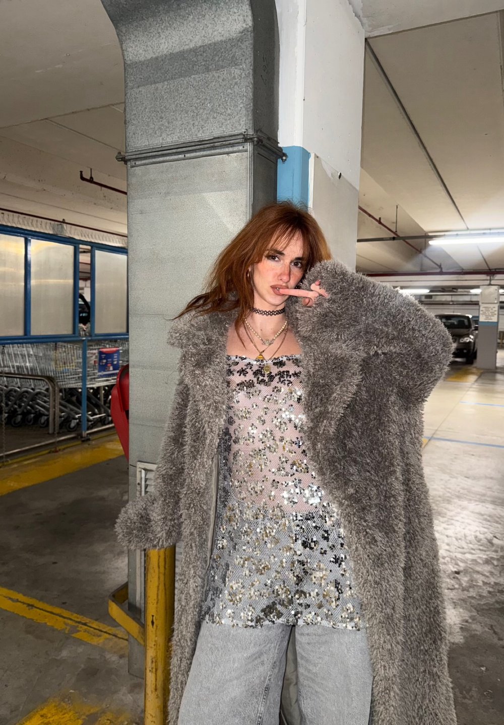 Manteau long gris en fausse fourrure, haut transparent à paillettes avec motifs floraux, jean baggy clair délavé, posant dans un parking.