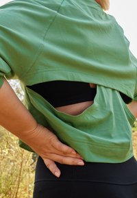 Camisa verde cropped com corte nas costas, sobrepondo uma camisola preta. Vista em close, textura suave, mangas curtas e detalhes de costura.