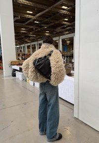 Personne portant une veste beige duveteuse, un jean large bleu, des chaussures noires et un sac à dos en cuir noir dans un marché couvert.