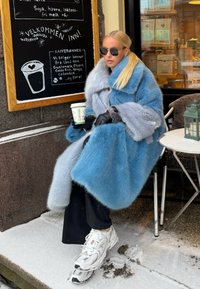 Ανοιχτό μπλε παλτό γούνας με γκρίζες λεπτομέρειες, μαύρα γάντια, μαύρο παντελόνι και λευκά αθλητικά παπούτσια. Καθισμένος σε καφετέρια με ένα φλιτζάνι καφέ πάνω σε χιονισμένη επιφάνεια.