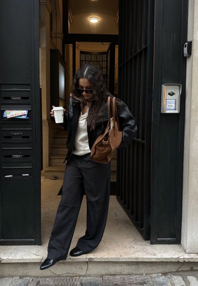 Mujer con gafas de sol, chaqueta de cuero negra y pantalones de rayas bajando la entrada de un edificio mientras sostiene una taza de café y un bolso marrón.